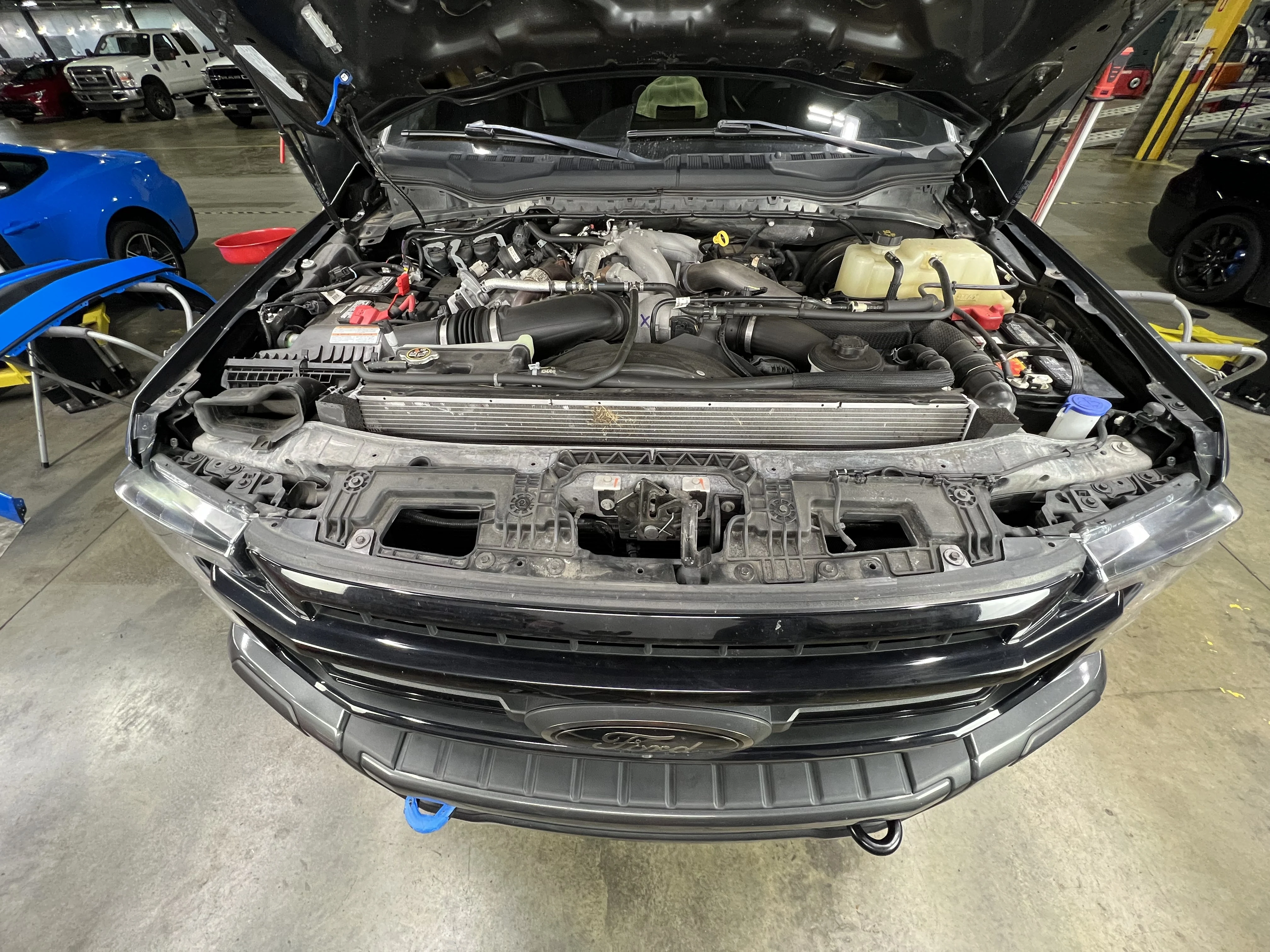 The engine bay of a Ford F-250 Super Duty 6.7L Powerstroke with the hood open. Just out of frame there is a blue Ford Mustang to the left and black Ford Mustang GT to the right.