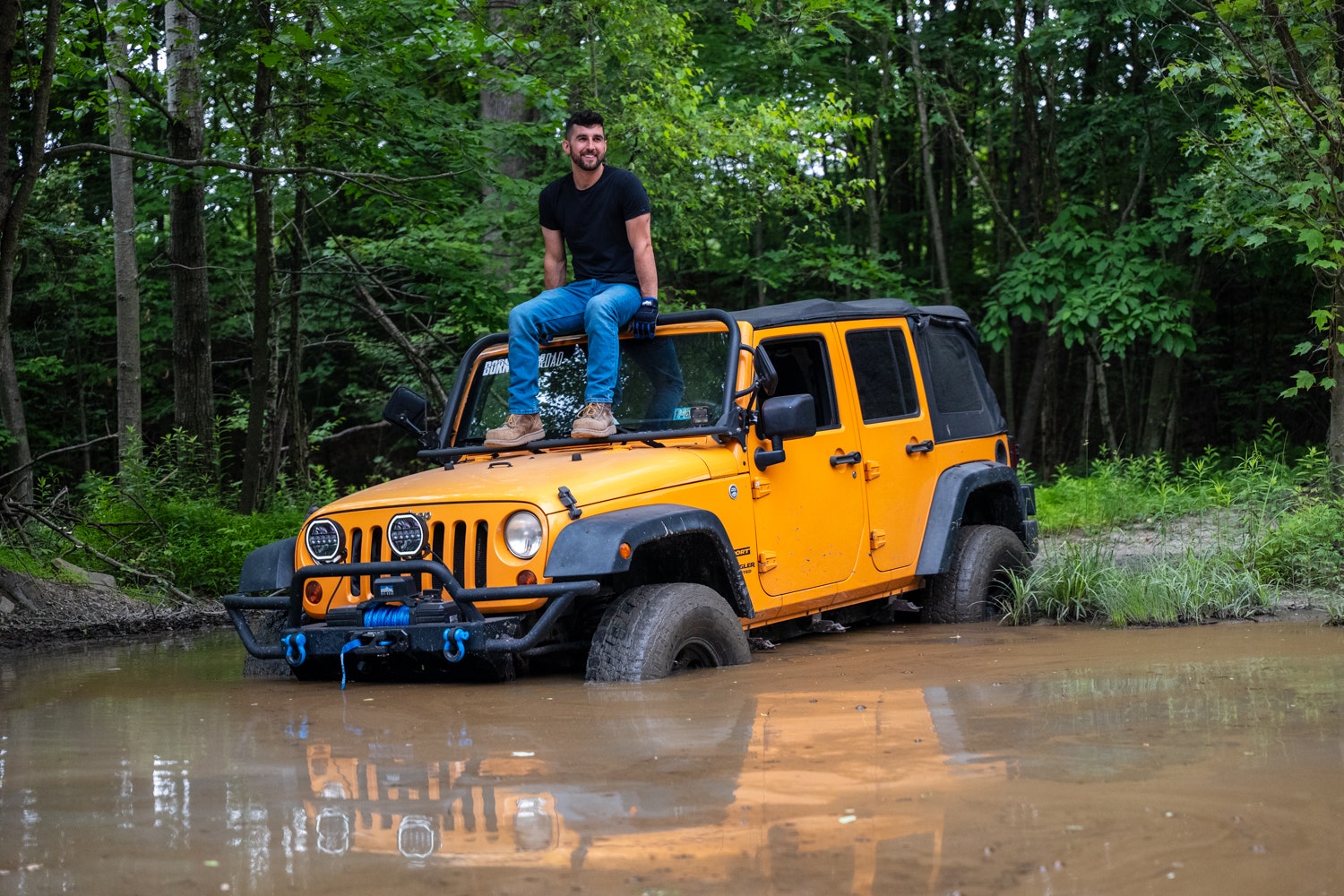 An image of Anthony, a Mishimoto engineering, sitting on the hood of a yellow Jeep JK Wrangler that is in the middle of a mud bog.